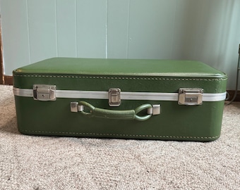 May include: A rectangular olive green suitcase featuring a smooth exterior finish, a central handle, and three silver-toned metal latches along the front edge. The suitcase is constructed with a reinforced frame and visible stitching details.