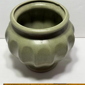 May include: A sage green ceramic pot with a textured surface and a scalloped design. The pot has a wide opening and sits on a pedestal base. The pot measures approximately 5 inches in diameter.