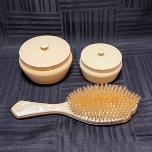 May include: A vintage vanity set featuring two small, lidded jars and a hairbrush. The jars are a light beige color with a round shape. The hairbrush has a light beige handle with a decorative design and natural bristles.