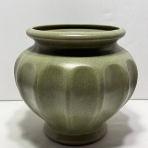 May include: A sage green ceramic vase with a rounded body and a flared rim. The vase has vertical, rounded indentations around its circumference. The base is a smaller, circular pedestal. The vase is approximately 18 cm tall.