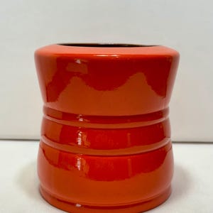 May include: A bright orange ceramic vessel with a glossy finish, featuring a cylindrical shape with two distinct horizontal ridges encircling the body and a wide, open top with a dark interior rim, showcasing a smooth and reflective surface texture.