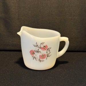 May include: A small, off-white creamer with a handle and a spout. The creamer features a floral design with pink flowers and green leaves. The creamer is made of ceramic and is likely vintage.