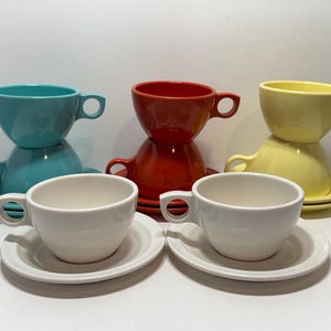 May include: A collection of colorful ceramic teacups and saucers. The cups are in shades of teal, red, yellow, and white. Each cup has a rounded shape with a handle. The saucers are round and white. The cups are stacked in pairs.