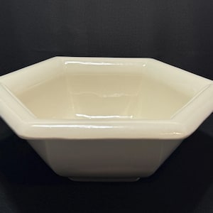 May include: Cream-colored, hexagonal ceramic bowl. The bowl has a smooth, glossy finish and a geometric design. The sides slope inward, and the rim is slightly rounded. A simple, elegant piece for serving or display.
