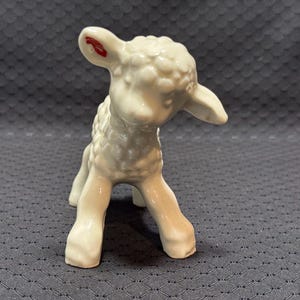 May include: A small, white ceramic figurine of a standing lamb with a textured, curly wool surface. The lamb has a glossy finish and a distinct red mark on the inner side of its left ear, with its head tilted slightly downward towards the ground.