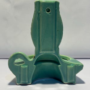 May include: A light green ceramic wall pocket with a glossy finish. The object features a vertical back panel with a small circular hole for hanging, a curved base, and a side loop handle. The design is symmetrical with smooth, molded edges.
