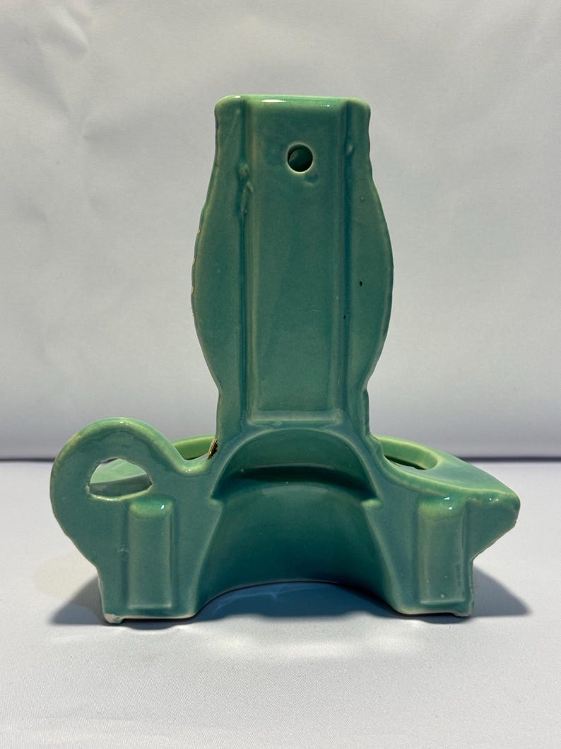 May include: A light green ceramic wall pocket with a glossy finish. The object features a vertical back panel with a small circular hole for hanging, a curved base, and a side loop handle. The design is symmetrical with smooth, molded edges.