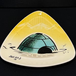 May include: Triangular ceramic ashtray with a yellow and white mountain scene. The center features a teal and black igloo design. The ashtray has the text "SASCHA B" on the front.