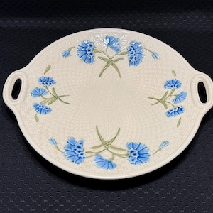 May include: A round, cream-colored ceramic serving platter with two integrated side handles. The surface features a raised basket-weave texture and is decorated with three clusters of blue cornflower blossoms with green stems and leaves.