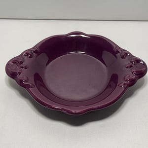 Op de afbeelding: Een dieppaarse keramische schaal met een geschulpte rand en decoratieve bloemaccenten. De schaal heeft een ondiep, rond interieur en wordt waarschijnlijk gebruikt om te serveren of tentoon te stellen. De kleur is een rijke, verzadigde tint.