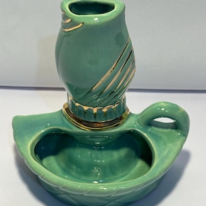 May include: A teal ceramic candle holder featuring a rounded base with a handle and a central cylindrical socket. The piece is decorated with gold-colored accents along the rim of the socket and diagonal gold stripes on the upper section.