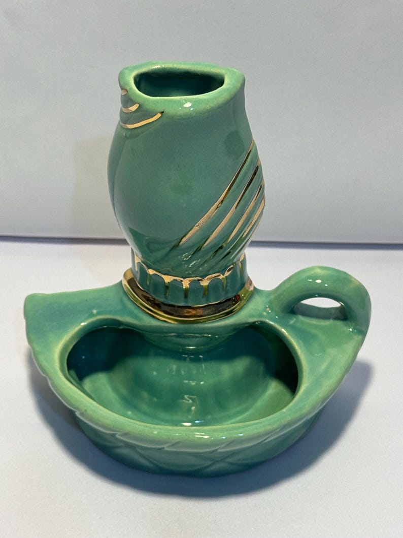May include: A teal ceramic candle holder featuring a rounded base with a handle and a central cylindrical socket. The piece is decorated with gold-colored accents along the rim of the socket and diagonal gold stripes on the upper section.