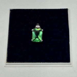May include: A rectangular, bright green gemstone pendant with a silver-colored setting. The pendant is displayed on a dark blue velvet surface inside a white-edged box. The gemstone is approximately 1 inch in length.