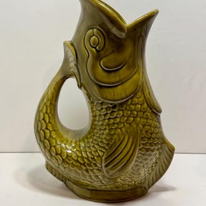 May include: A ceramic pitcher in the shape of a fish, with a handle that forms the fish's tail. The pitcher is a muted olive green color, with scale detailing covering the body. The mouth of the fish is open, forming the spout.