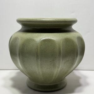 May include: A sage green ceramic vase with a rounded body and a scalloped design. The vase has a wide opening and a small, circular base. The surface has a speckled texture. This decorative vase is suitable for displaying flowers or as a standalone accent piece.