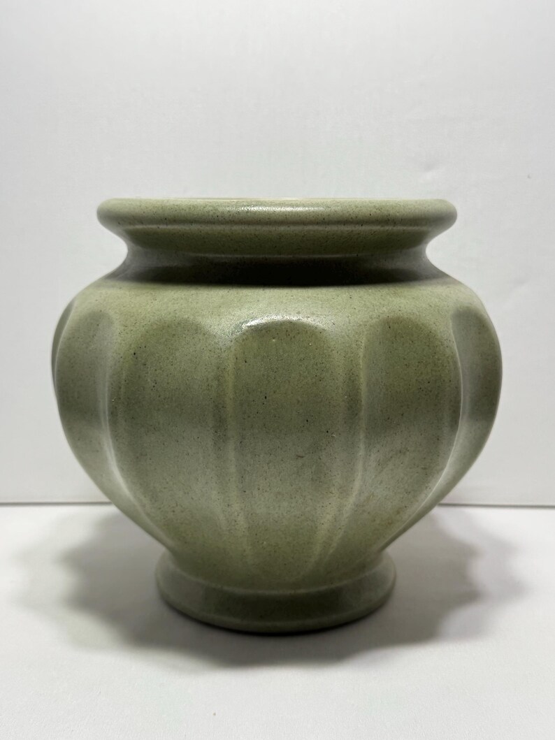May include: A sage green ceramic vase with a rounded body and a scalloped design. The vase has a wide opening and a small, circular base. The surface has a speckled texture. This decorative vase is suitable for displaying flowers or as a standalone accent piece.