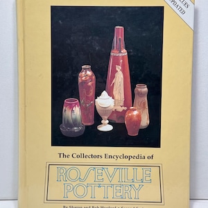 Puede incluir: Un libro de tapa dura titulado The Collectors Encyclopedia of Roseville Pottery, Second Series por Sharon y Bob Huxford. La cubierta de color amarillo pálido presenta un recuadro rectangular negro con varios jarrones de cerámica y un recipiente con tapa.