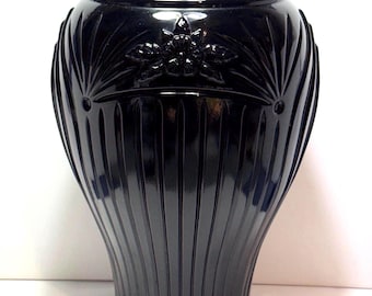 May include: A glossy black glass vase featuring a wide, rounded body that tapers towards the base and a flared rim. The surface is decorated with vertical fluted lines and a raised floral motif centred near the top, creating a textured appearance.