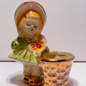 May include: A vintage ceramic figurine of a girl in a green dress and a hat, holding a basket. The figurine has gold accents on the hat, dress, and basket. The girl's eyes are closed, and she has a red bow.