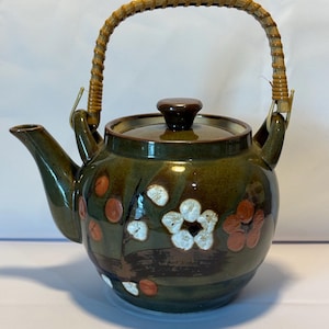 Puede incluir: Una tetera de cerámica verde con tapa marrón y asa de bambú trenzado. La tetera está decorada con flores blancas y naranjas.