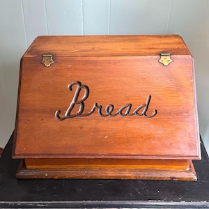 Può includere: Un portapane rettangolare in legno con coperchio frontale inclinato. La scatola presenta una calda finitura in venature del legno marrone e due cerniere in metallo color ottone sulla parte superiore. La parola "Bread" è scritta in corsivo metallico.