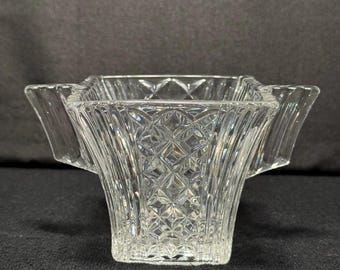 May include: A clear glass square bowl with a diamond pattern and flared sides. The bowl has two handles on opposite sides. The glass is transparent, allowing light to pass through, and is set against a dark background.