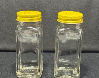 May include: Two clear glass shakers with square bases and bright yellow screw-on metal lids. Each lid features a series of small holes for dispensing contents. The glass bodies are transparent and rectangular in shape, standing side by side.
