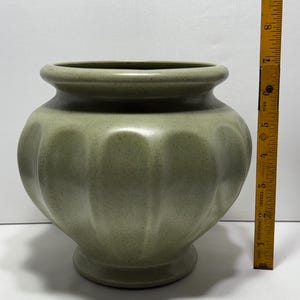 May include: A sage green ceramic vase with a rounded body and a flared rim. The vase has a ribbed design and sits on a small, circular base. The vase is approximately 7 inches tall.