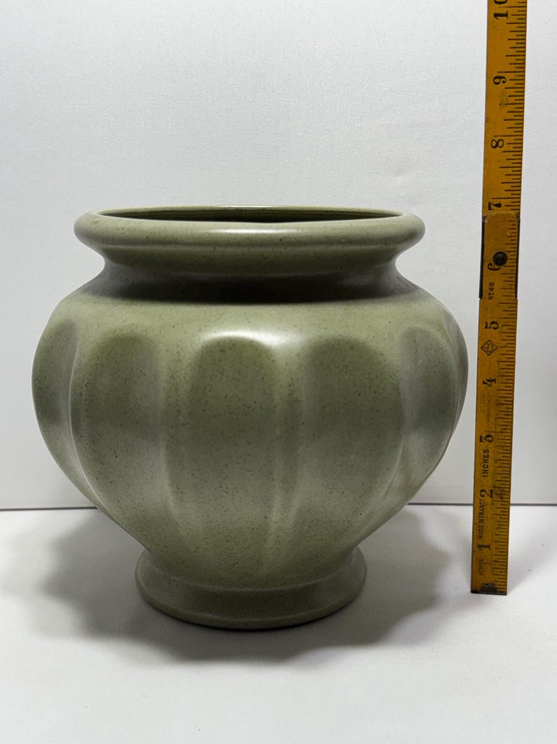 May include: A sage green ceramic vase with a rounded body and a flared rim. The vase has a ribbed design and sits on a small, circular base. The vase is approximately 7 inches tall.