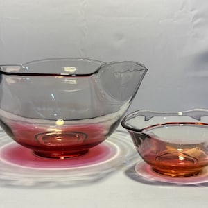 May include: Two clear glass bowls with a red-orange base and rim. The larger bowl has a spout and a wavy rim. The smaller bowl has a similar design. The bowls are sitting on a white surface.