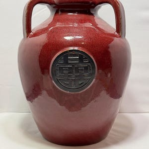 Puede incluir: Un jarrón de cerámica rojo grande con dos asas y un emblema negro. El jarrón tiene un acabado brillante y una forma redondeada. El emblema es un diseño circular con un patrón geométrico.