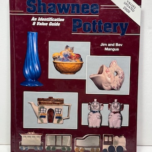 Peut inclure: Un livre à couverture rigide intitulé Shawnee Pottery: An Identification & Value Guide par Jim et Bev Mangus. La couverture rouge foncé présente des images de divers objets en céramique, dont un vase bleu, un bol de fruits et un train.