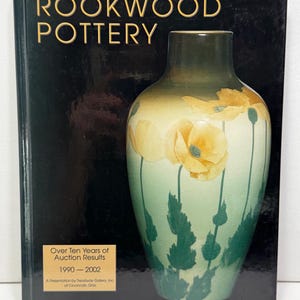 May include: A hardcover book titled Rookwood Pottery with a black cover featuring a large vase illustration. The vase has a green to cream gradient with yellow poppy flowers and green stems. A gold label reads Over Ten Years of Auction Results 1990-2002.