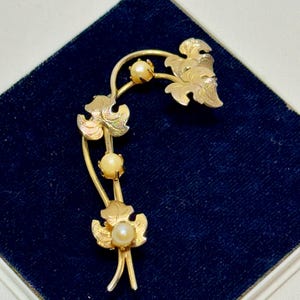 May include: A gold-toned brooch featuring a stylized floral design with three faux pearls. The brooch has a curved shape with detailed leaf and stem elements. The brooch is displayed on a dark blue velvet surface.