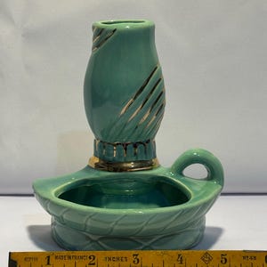 May include: A teal ceramic candle holder with a gold-trimmed base and a vertical gold-accented neck. The holder features a small handle and a wide, shallow bowl. A yellow measuring tape placed in front shows the object is approximately 5 inches wide.