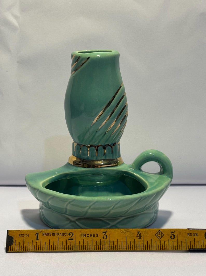May include: A teal ceramic candle holder with a gold-trimmed base and a vertical gold-accented neck. The holder features a small handle and a wide, shallow bowl. A yellow measuring tape placed in front shows the object is approximately 5 inches wide.