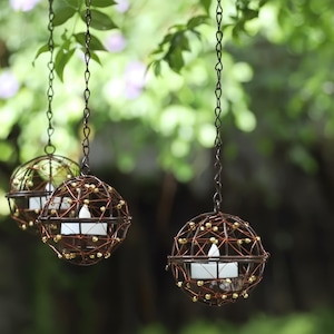 Puede incluir: Tres candelabros esféricos colgantes. Cada soporte está hecho de una jaula de metal marrón oscuro, adornada con pequeñas cuentas doradas. Una vela blanca se encuentra dentro de cada esfera, suspendida por una cadena contra un fondo verde borroso.