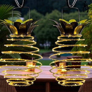 Puede incluir: Dos luces solares colgantes con forma de piña, con diseños en espiral y luces LED blancas cálidas. Las luces tienen una parte superior de metal con forma de piña con detalles verdes y dorados. Cuelgan de un gancho de metal.
