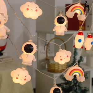 Könnte beinhalten: Eine LED-Lichterkette mit weißen lächelnden Wolken, weißen Astronauten mit schwarzen Helmen, bunten Regenbögen und weißen Raketen mit roten und blauen Akzenten. Die dekorativen Lichter sind mit einem dünnen, flexiblen Draht verbunden.