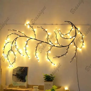 Op de afbeelding: Een decoratieve wandlamp in de vorm van een uitgestrekte boomtak met dunne, donkere en flexibele stelen. De tak is voorzien van talloze kleine, warmwitte LED-lampjes aan de uiteinden van de twijgen, wat een gloeiend, organisch silhouet creëert.
