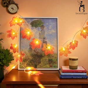 May include: A decorative string of orange and yellow maple leaf lights, with a painting and a candle on a wooden surface. The lights are draped over the painting. A small clock is also visible.