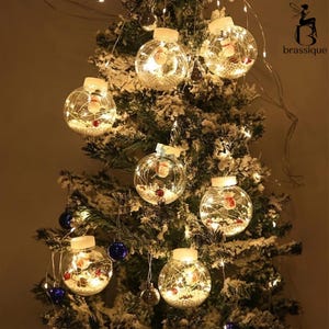 以下が含まれることがあります： 雪だるまのフィギュア、白いビーズ、ストリングライトが入った透明な丸いオーナメントで飾られたクリスマスツリー。オーナメントは照らされ、暖かくお祝いの輝きを作り出しています。青と金のオーナメントも表示されます。