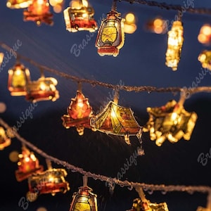 Pode incluir: Fio de luzes decorativas com tema de acampamento. O fio apresenta pequenos recortes iluminados de uma lanterna, uma fogueira, uma tenda e uma caravana. As luzes são enfiadas em um fio fino e torcido, com um fundo escuro.