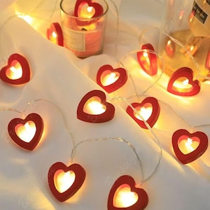 May include: A string of red heart-shaped LED lights with warm white illumination. Each heart features a hollow centre, and the lights are arranged on a thin, flexible wire. A glass candle holder with a red candle is visible in the background.