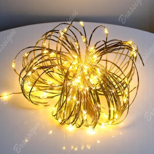 Może przedstawiać: Pęk elastycznych lampek typu fairy lights na miedzianym drucie, ułożonych w kształt kuli. Cienkie metalowe przewody są ze sobą splecione i wyposażone w liczne małe, ciepłe żółte diody LED, które emitują jasne światło w całej strukturze.