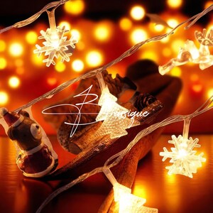 May include: A string of LED fairy lights featuring clear plastic snowflake and Christmas tree shapes, along with a small bear figurine wearing a red and white Santa hat. The lights emit a warm glow, connected by a thin, transparent wire.