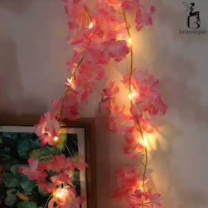 Puede incluir: Guirnalda de flores artificiales rosas con pequeñas luces LED blancas cálidas. La guirnalda cuelga de una pared, con una imagen enmarcada visible en la esquina inferior izquierda. El logotipo de Brassique está en la esquina superior derecha.
