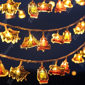 May include: String lights featuring camping-themed shapes: lanterns, tents, campfires, and RVs. The lights are warm yellow and red, strung on a brown wire, creating a cozy, outdoorsy ambiance. Perfect for decorating a camper or adding a touch of nature to any space.