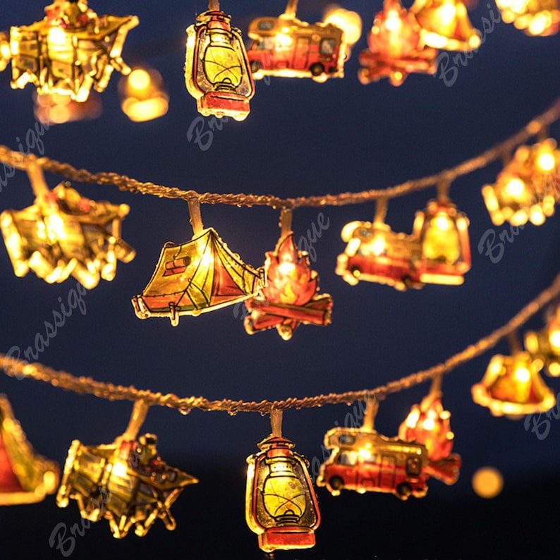 May include: String lights featuring camping-themed shapes: lanterns, tents, campfires, and RVs. The lights are warm yellow and red, strung on a brown wire, creating a cozy, outdoorsy ambiance. Perfect for decorating a camper or adding a touch of nature to any space.