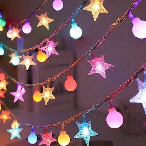 Puede incluir: Una guirnalda de luces LED decorativas con una alternancia de bombillas translúcidas en forma de estrella y bombillas esféricas sólidas. Las luces emiten un brillo multicolor en tonos rosa, azul, amarillo y blanco, unidas por un cable cobrizo.